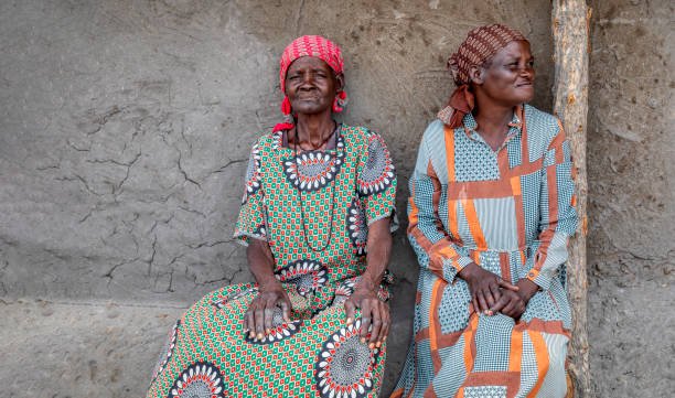 two elderly woman sitting next to the house wall in the yard in an African village